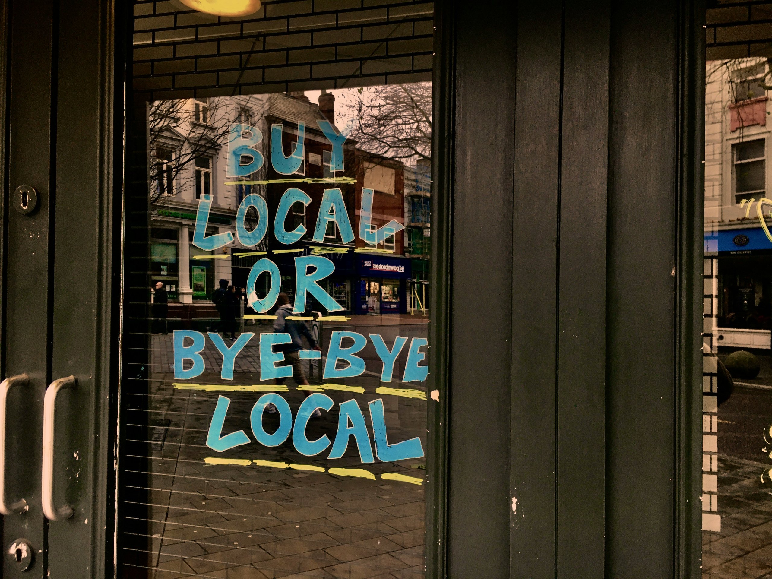 Glass door with blue and yellow painted text reading 'BUY LOCAL OR BYE-BYE LOCAL' and a street reflected in the glass.