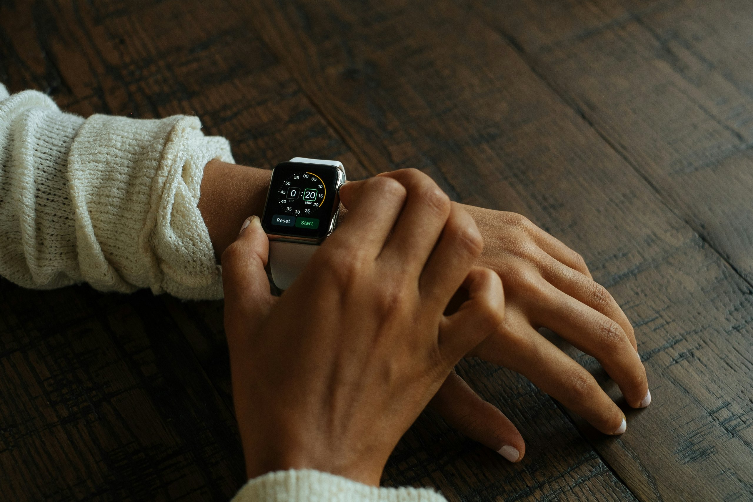 Person adjusting a white smartwatch timer set to 20 minutes while wearing a cream knitted sweater on a wooden surface.