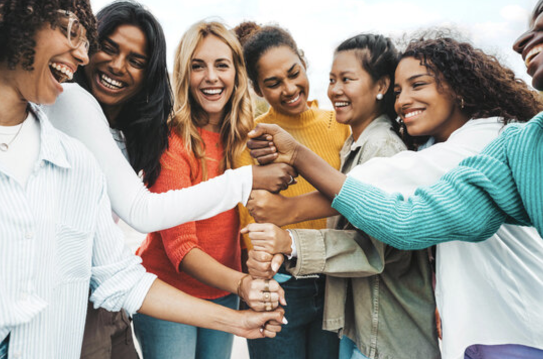 A diverse group of smiling women standing in a circle outdoors, stacking their hands together in a show of unity.