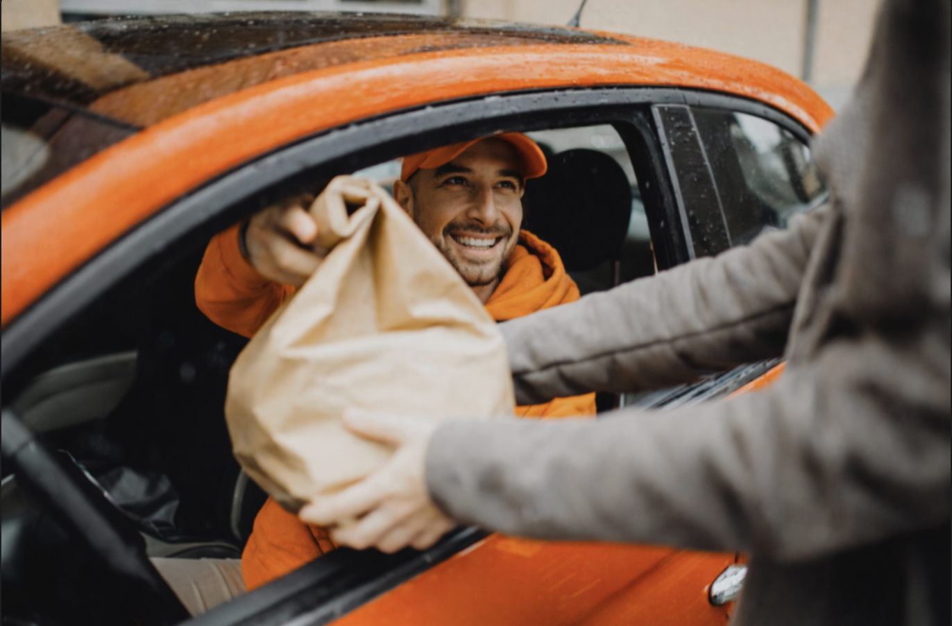 Smiling delivery person in orange outfit handing a brown paper bag to a customer through a car window.