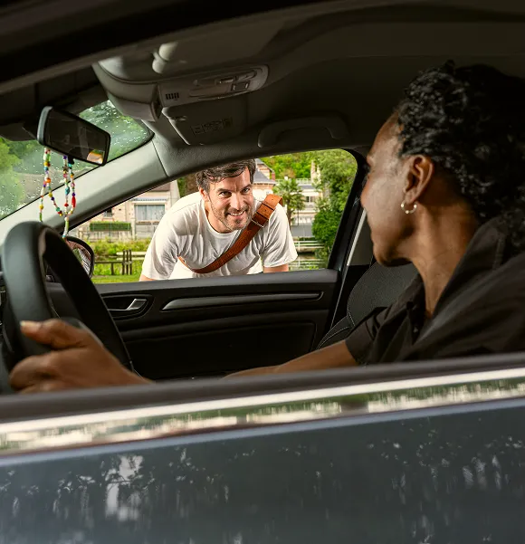 Man smiling and leaning toward the driver's side window of a car where a woman is sitting and looking at him.