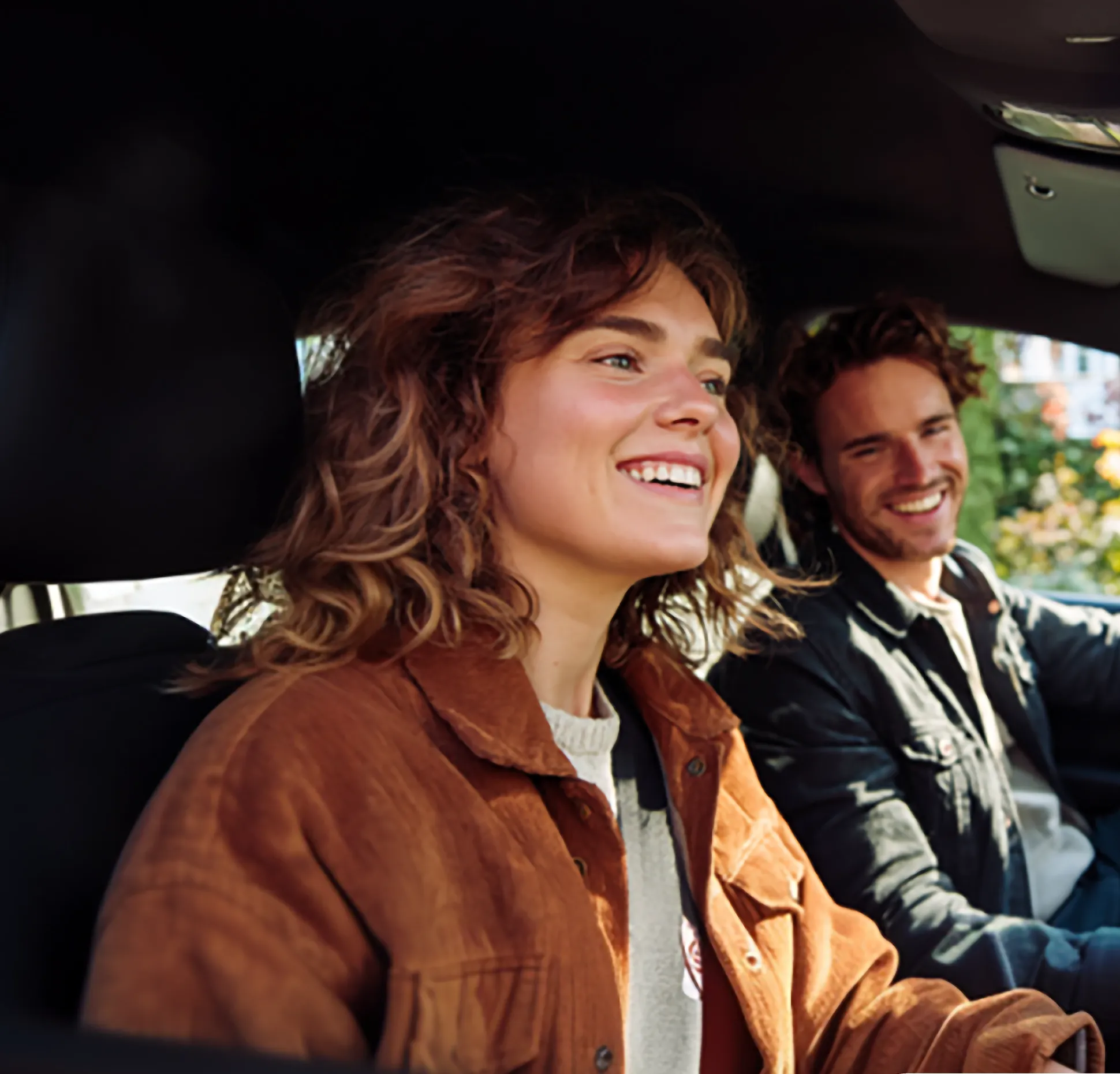 Smiling young woman and man sitting inside a car, with the man in the driver's seat and the woman in the front passenger seat.