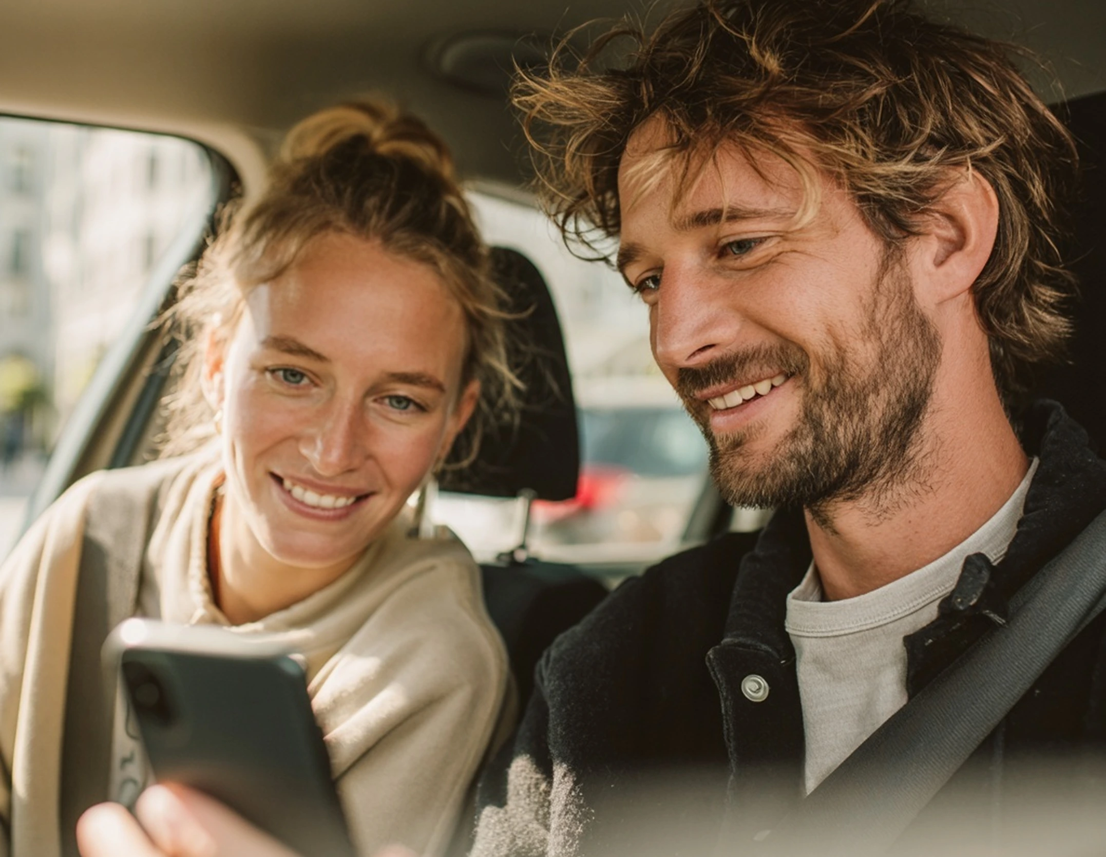 Smiling man and woman sitting in car looking at a smartphone.