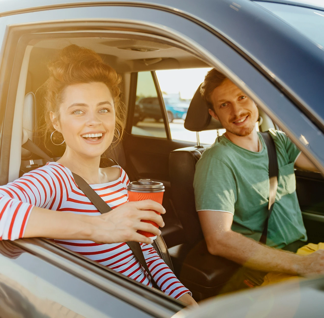 Smiling woman holding coffee cup in passenger seat and man in green shirt driving a car.