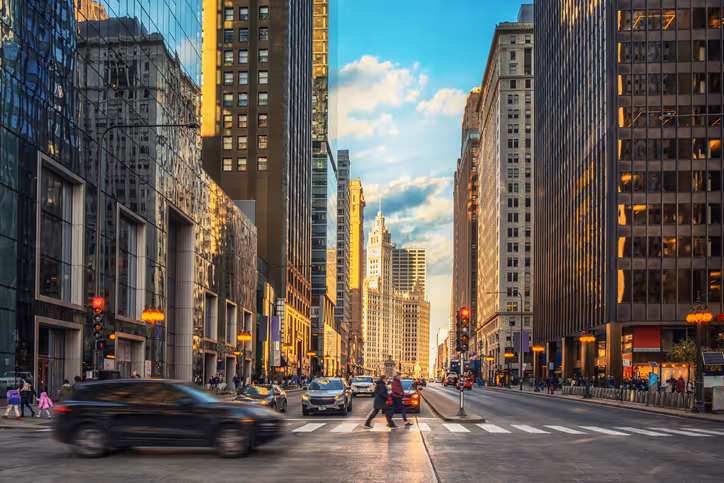 A busy city street captured at dusk, with pedestrians crossing at a crosswalk. Tall buildings line the road, reflecting warm sunlight, and vehicles are seen moving in both directions. A clear blue sky with scattered clouds adds depth to the scene.