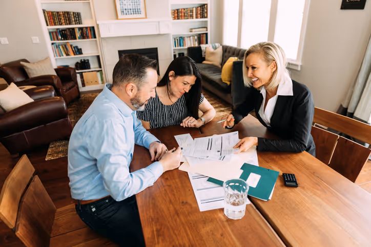 Three professionals engaged in a discussion around a wooden table, reviewing documents. A man in a light blue shirt, a woman in a striped top, and a woman in a black suit are collaborating in a cozy, well-lit living room.