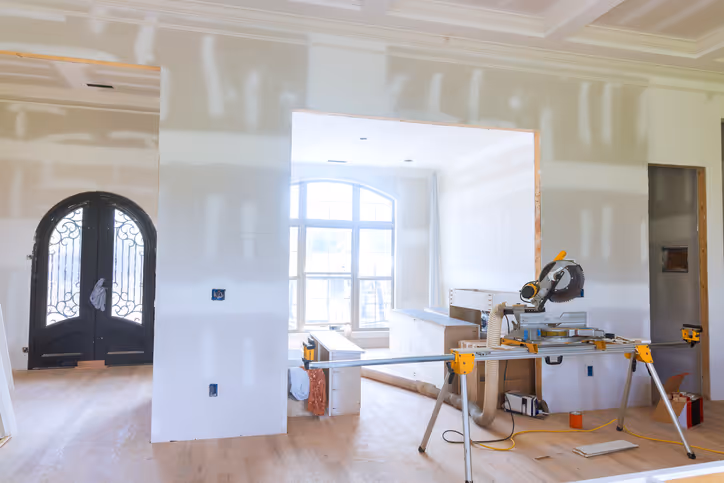 An unfinished interior space with drywall awaiting final touches. A large window lets in natural light, and a miter saw is positioned on a stand. Elegant double doors are visible on the left, showcasing the ongoing construction work.