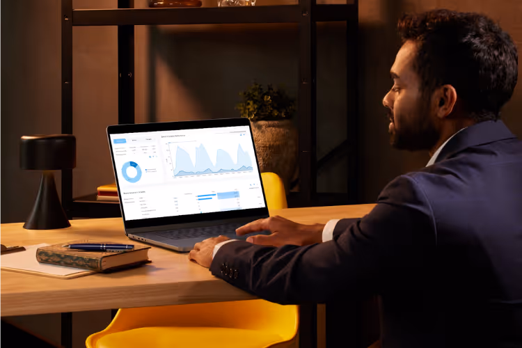 A man in a suit sitting at a desk with a laptop displaying data graphs and analytics. A notebook and pen are on the desk, alongside a black lamp in a well-lit room.