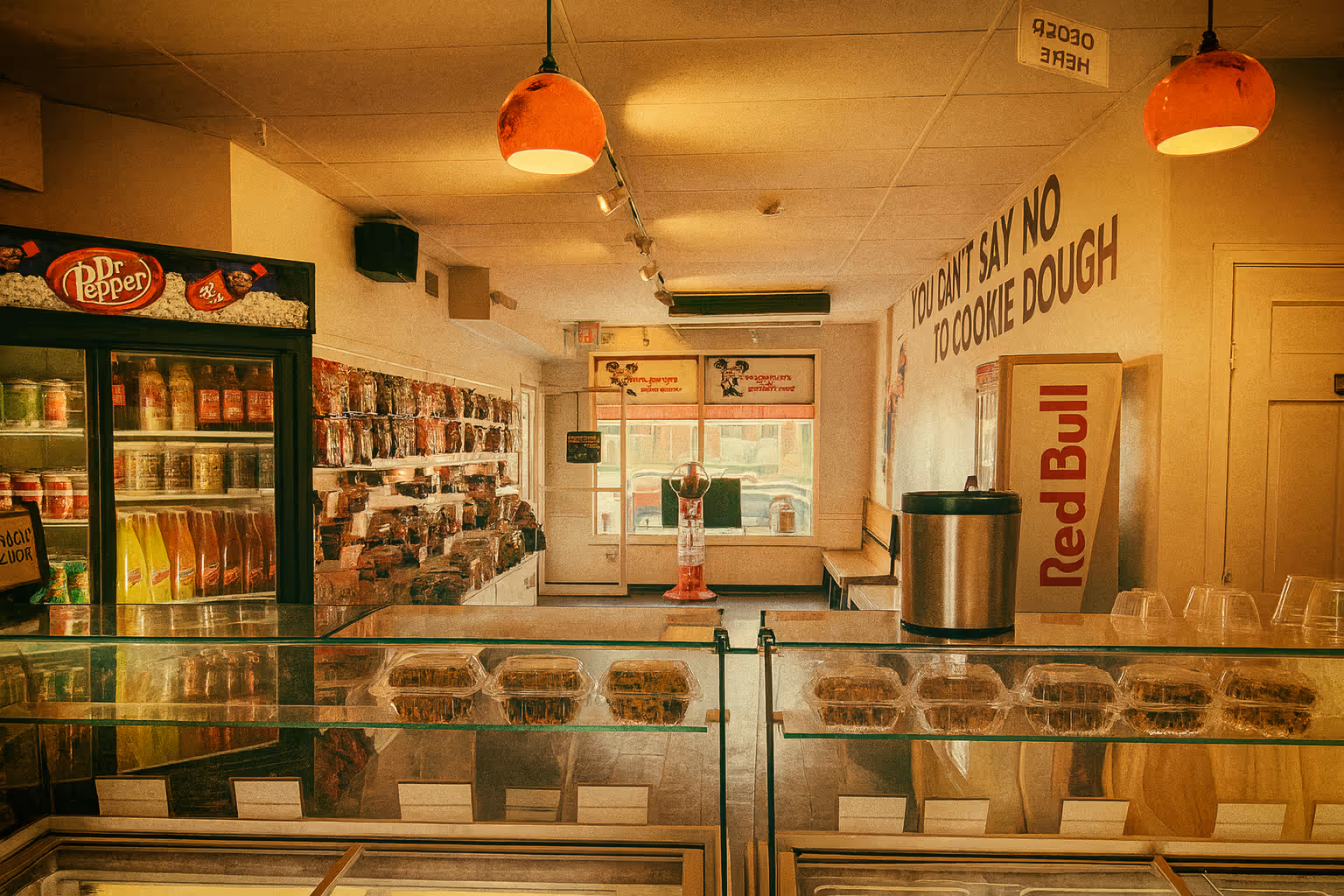 A brightly lit interior of a dessert shop, featuring a glass display case filled with treats. Refrigerators on the left hold various drinks, and playful signage on the wall says, 'YOU CAN'T SAY NO TO COOKIE DOUGH.'