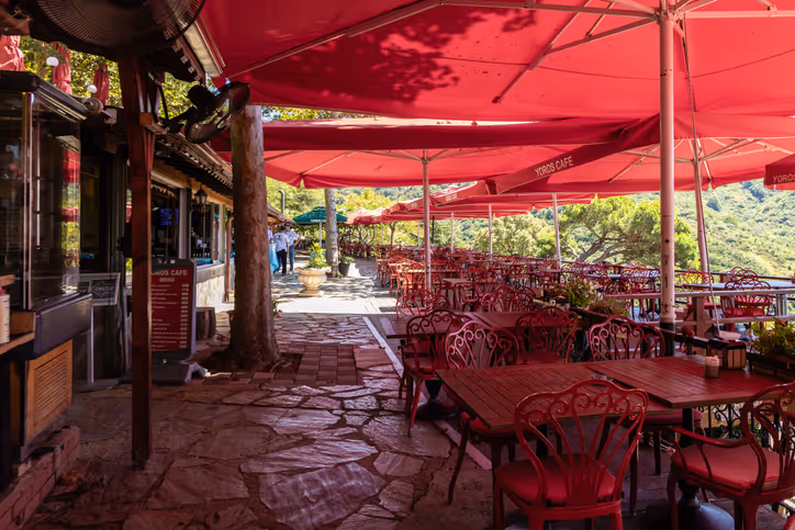 An outdoor café with red umbrellas providing shade over numerous red metal tables and chairs, situated on a stone pathway surrounded by greenery and hills in the background, creating a relaxing dining atmosphere.