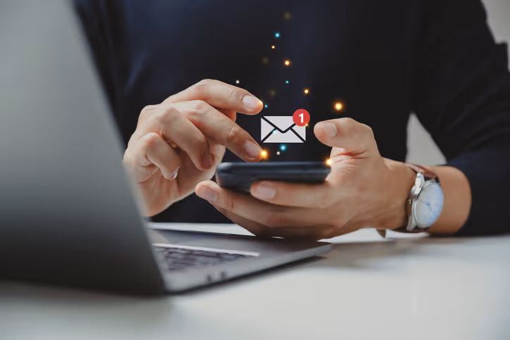 A person holding a smartphone with an envelope icon and notification badge, indicating a new message. A laptop is visible in the background on a white desk.