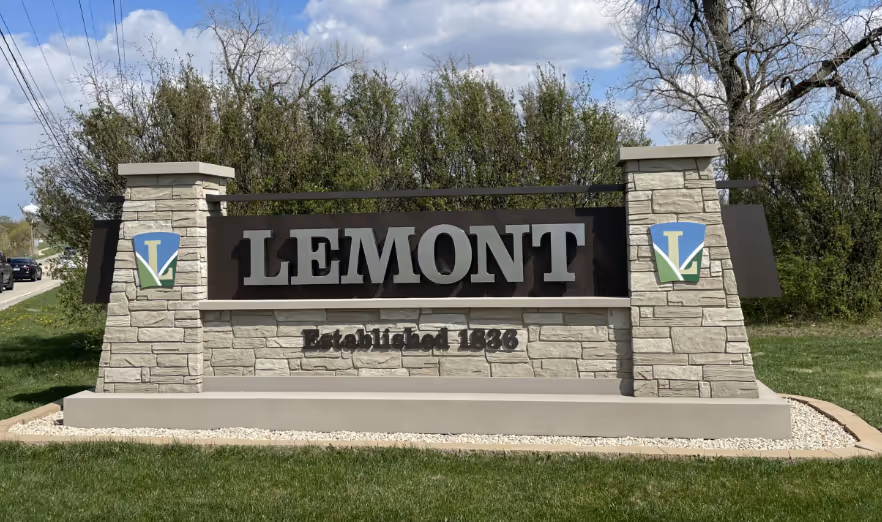 Welcome sign for Lemont, Illinois, featuring large black letters spelling "LEMONT" with the establishment year "Established 1888" below. The sign is flanked by stone pillars and greenery in the background under a partly cloudy sky.