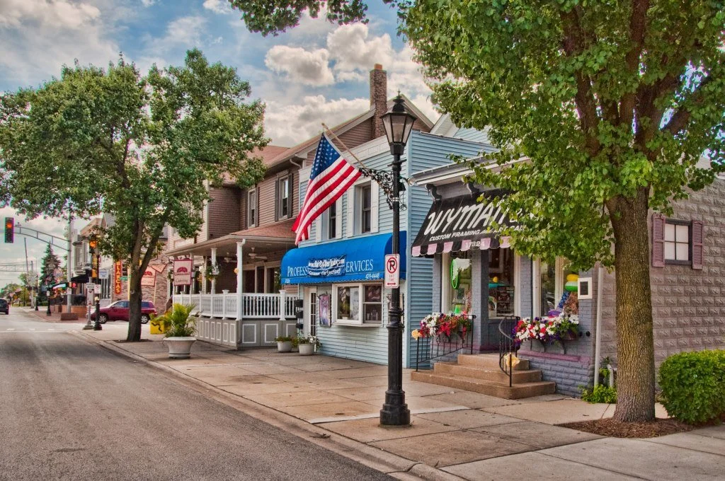 A quaint street scene featuring small shops with colorful awnings and an American flag hanging from a lamppost. Lush green trees line the sidewalk, and a clear sky fills the background, creating a charming small-town atmosphere.