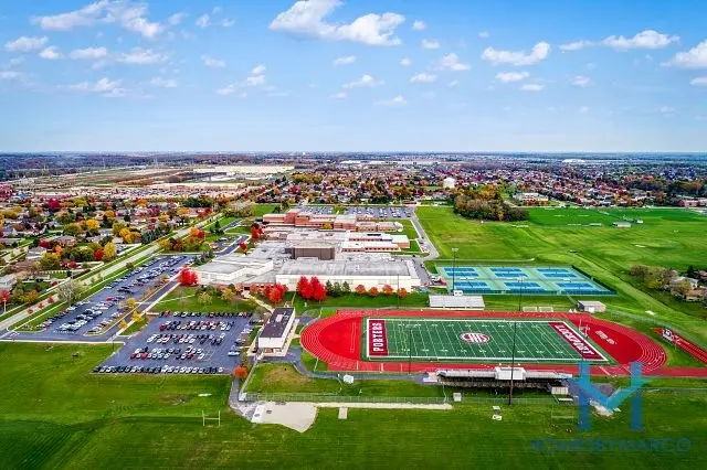 Aerial view of a school campus featuring sports fields, tennis courts, and ample parking, bordered by green fields and a suburban area. The school's red athletic field is prominently visible, surrounded by trees showing autumn colors.
