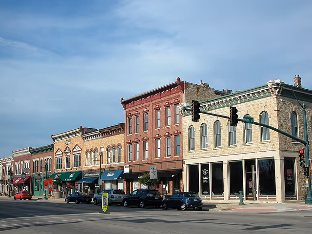 A view of a charming main street featuring a row of historic brick buildings with colorful facades. Various shops and storefronts line the street, and parked cars are visible along the curb under a partly cloudy sky.