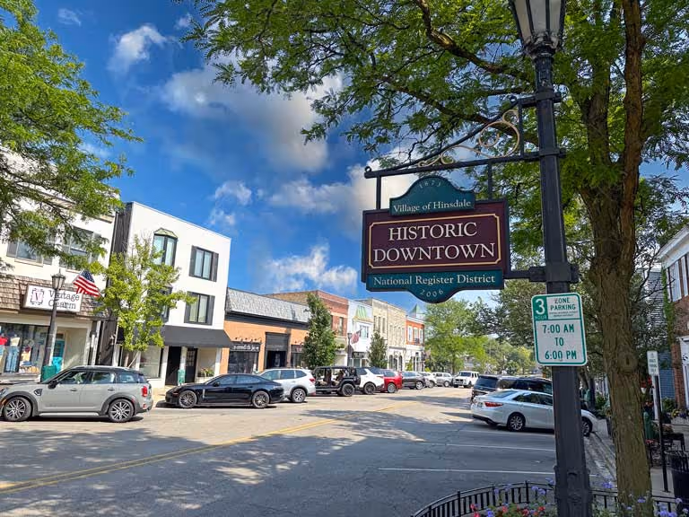 A view of a tree-lined street in a small downtown area, featuring a large sign that reads "Historic Downtown". Storefronts line the road, with parked cars and blue sky framing the scene.