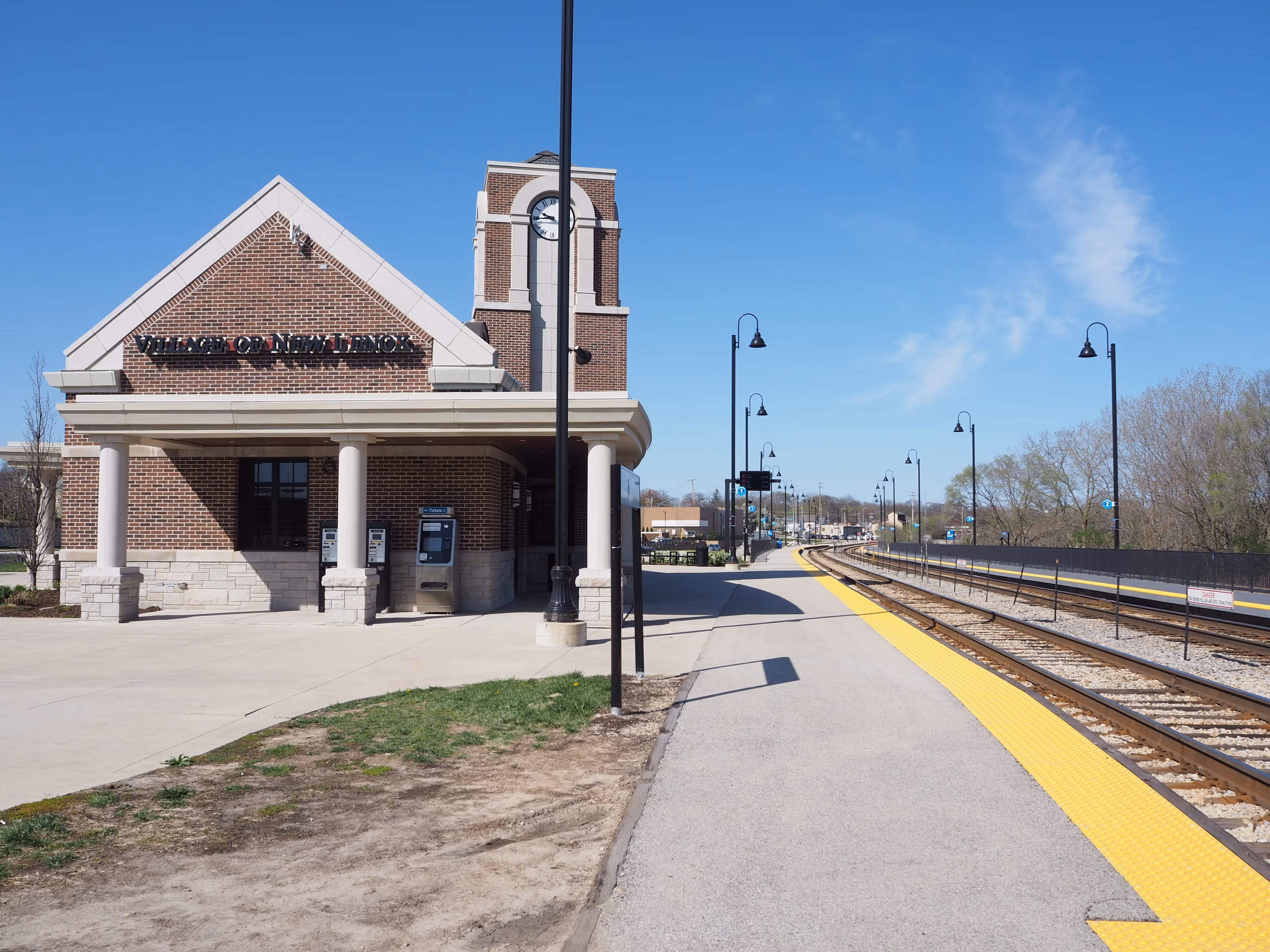 A view of a train station building featuring brick façade and a clock tower, with a yellow platform beside the railroad tracks. Street lamps line the pathway, and trees are visible in the background under a clear blue sky.