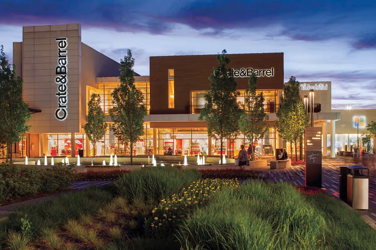 The exterior of a Crate & Barrel store at dusk, featuring a modern design with large windows. Surrounding the building are landscaped green areas with shrubs and flowers, and a softly illuminated water feature is visible in the foreground.