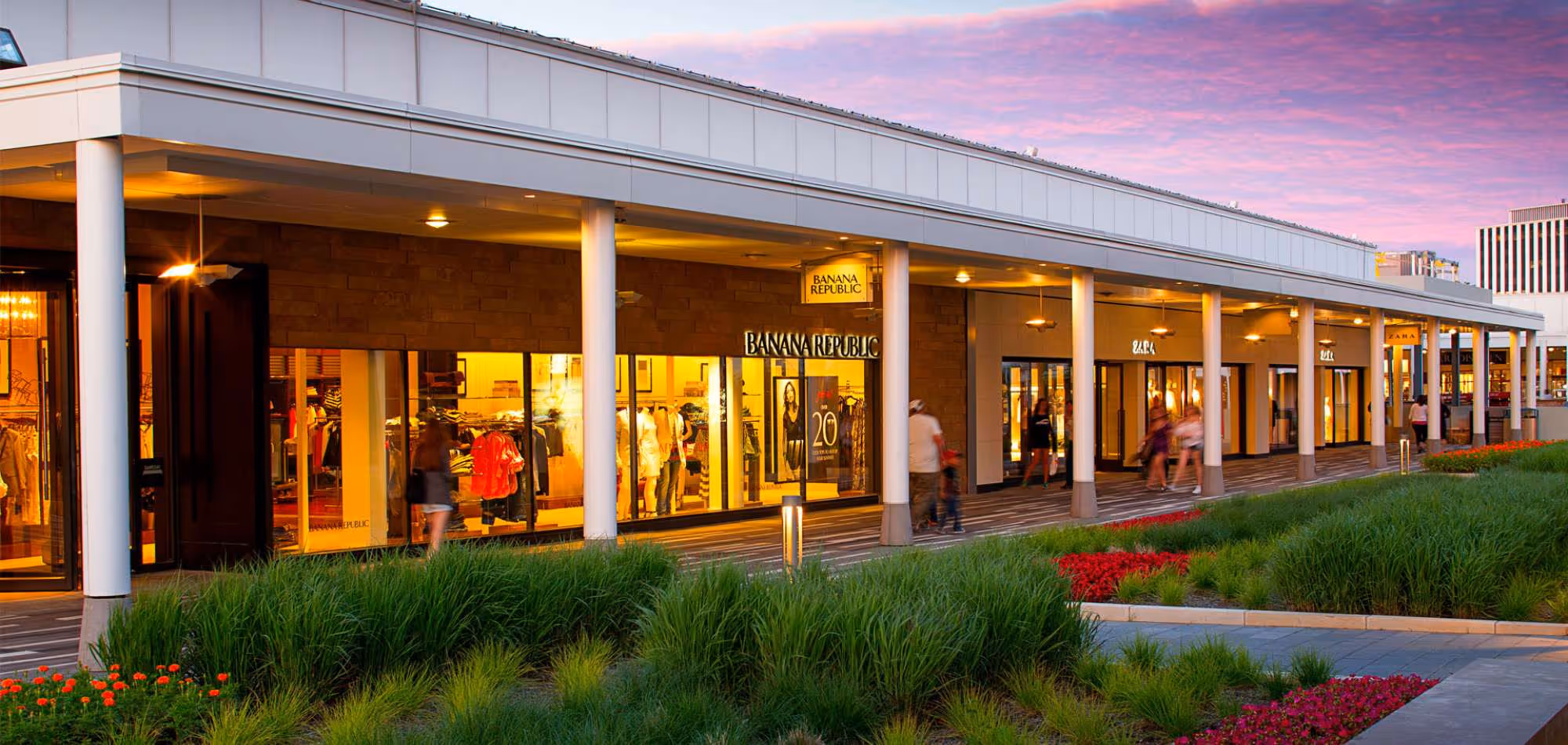 A view of a modern shopping plaza at sunset, featuring various storefronts with brightly lit displays. Shoppers stroll along the pathway, surrounded by landscaped greenery and colorful flower beds.