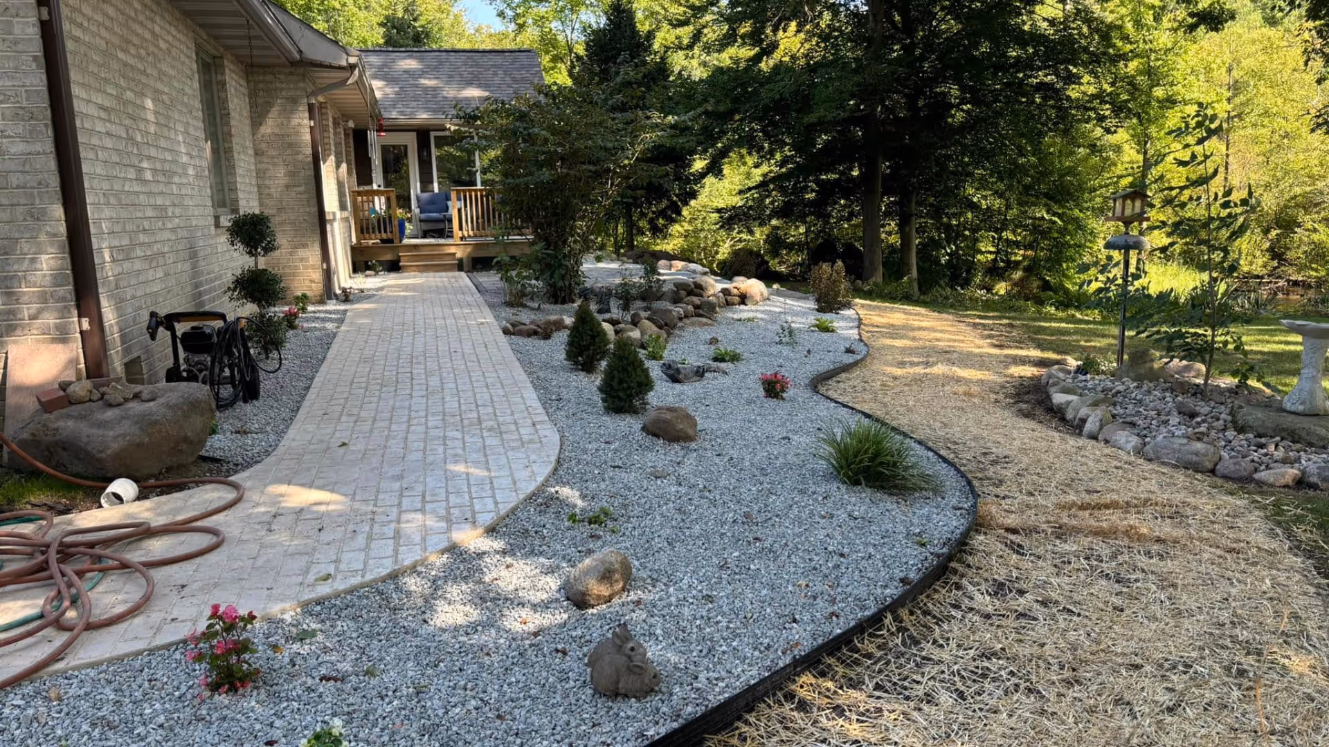 Curved stone walkway alongside a house with a landscaped garden area of white gravel, small shrubs, rocks, a birdhouse, and a mulched path surrounded by trees.