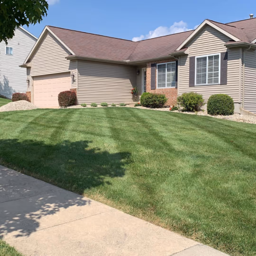 Single-story house with beige siding, brown roof, manicured lawn with striped mowing pattern, and a concrete driveway.
