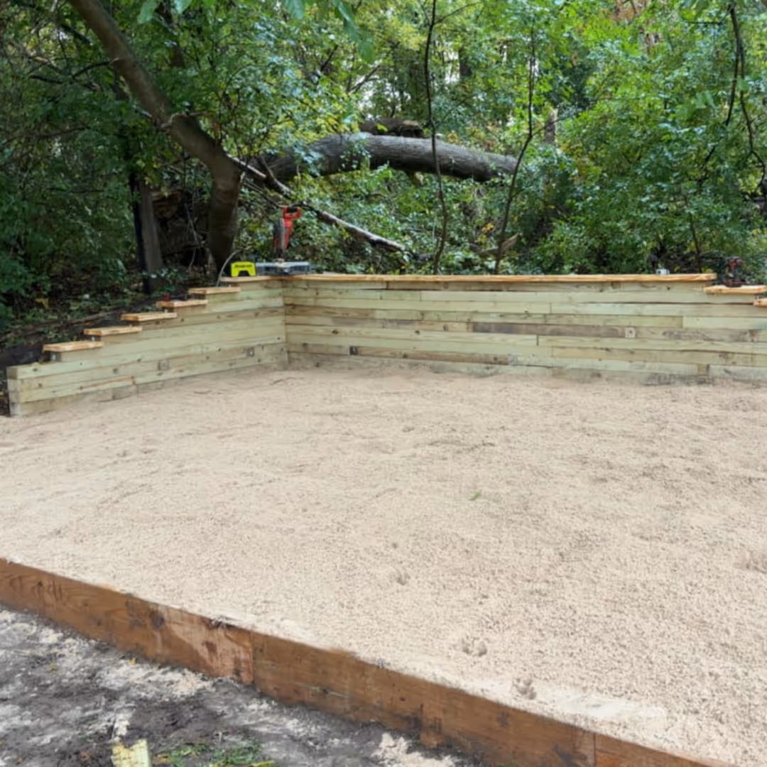 A rectangular sandbox with wooden walls and a smooth sand base, set in a forested area with dense green trees.