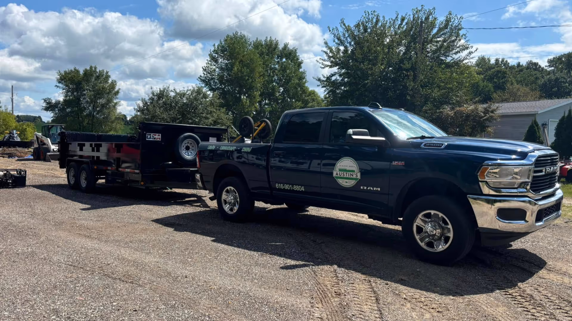 Blue RAM 2500 truck towing a black trailer with equipment, parked on a gravel lot with trees and a building in the background.