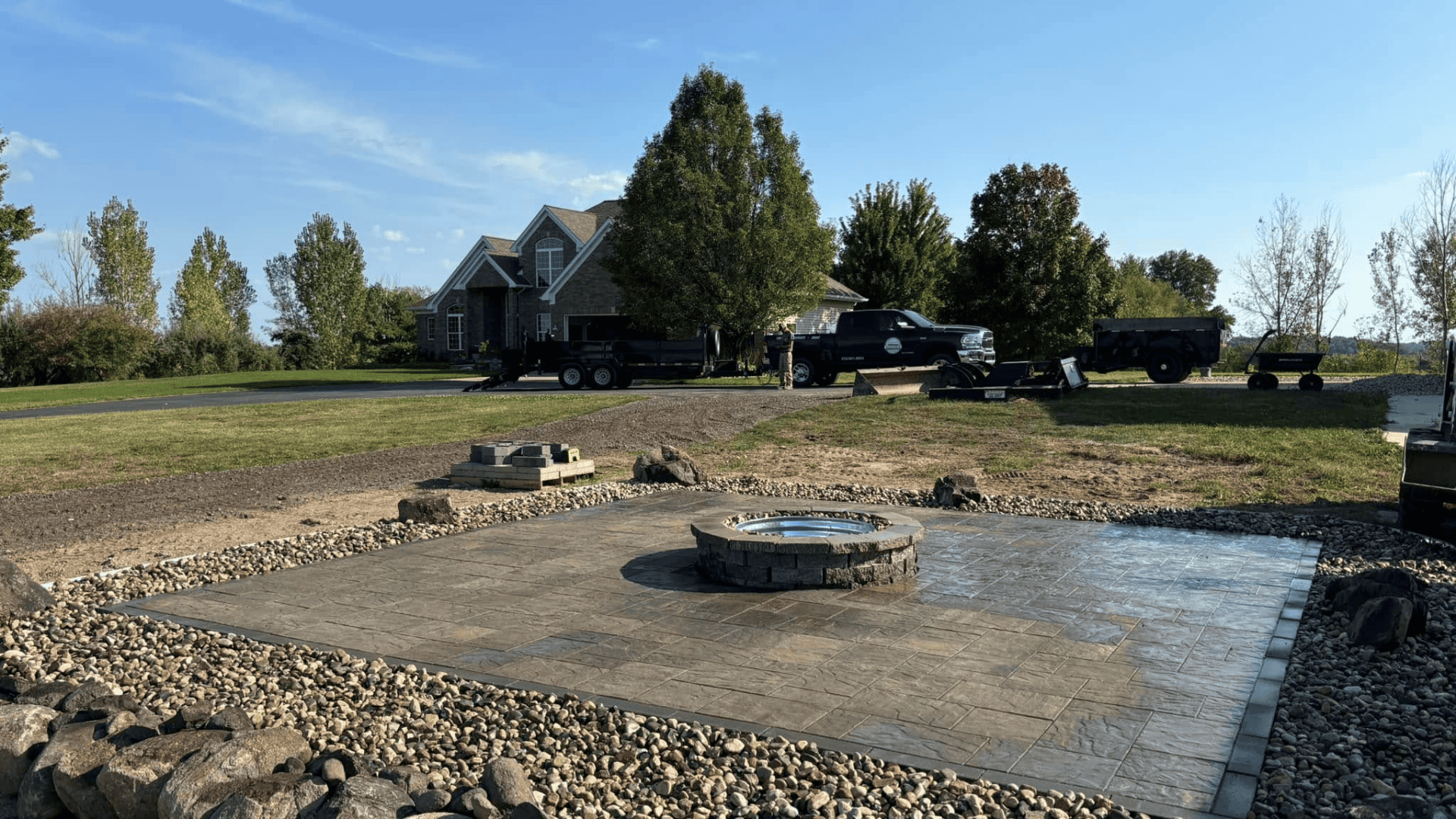 Patio with stone fire pit surrounded by pebbles in front of a suburban house with trucks and landscaping equipment nearby.