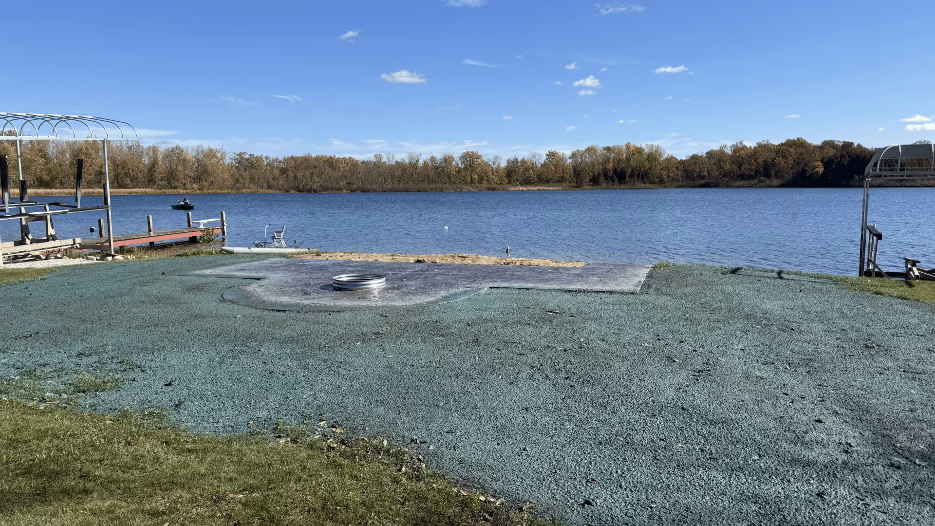 View of a calm lake with autumn trees in the background and a grassy shore in the foreground under a clear blue sky.