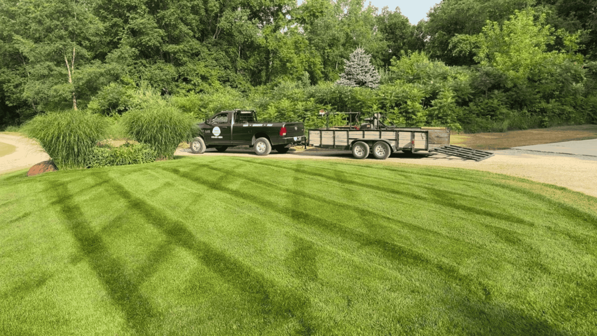 Black pickup truck with attached trailer parked on a gravel path beside a freshly mowed lawn and dense green trees.