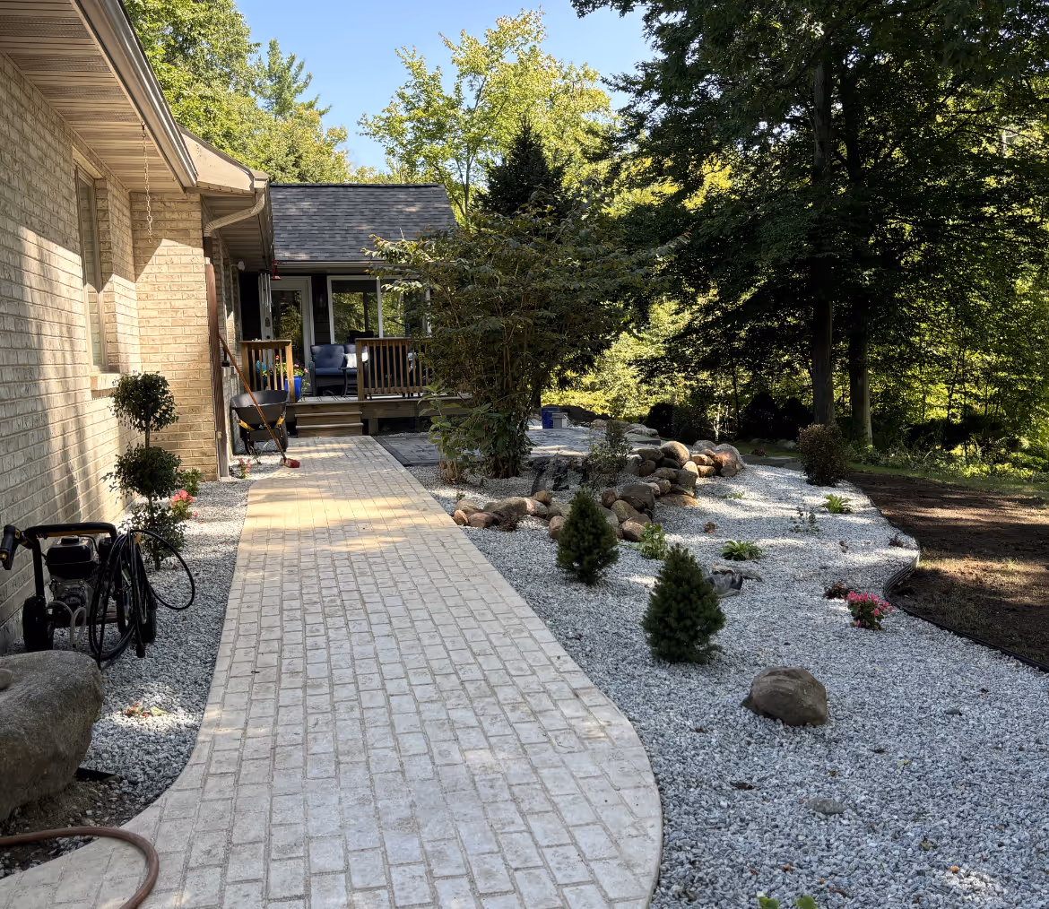 Brick pathway leading to a house porch with blue chairs, surrounded by gravel landscaping, trees, and rocks.