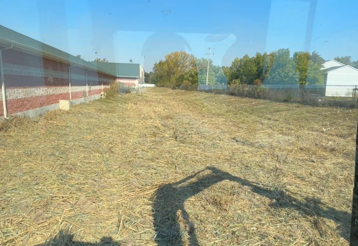 Open dry field next to a red and white building with trees and houses in the background under a clear blue sky.