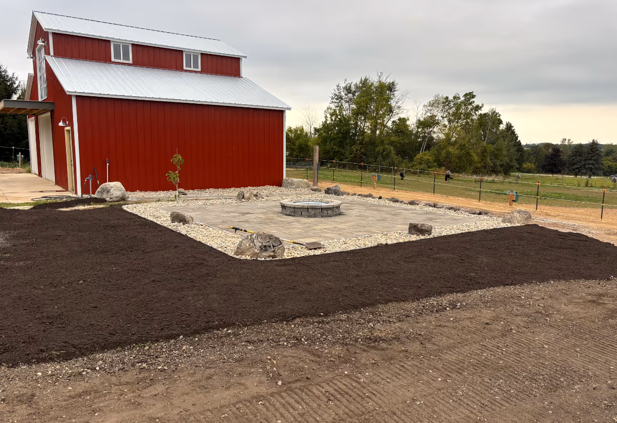 Red barn with a silver roof next to a square paved fire pit area bordered by white rocks and surrounded by dark soil under a cloudy sky.