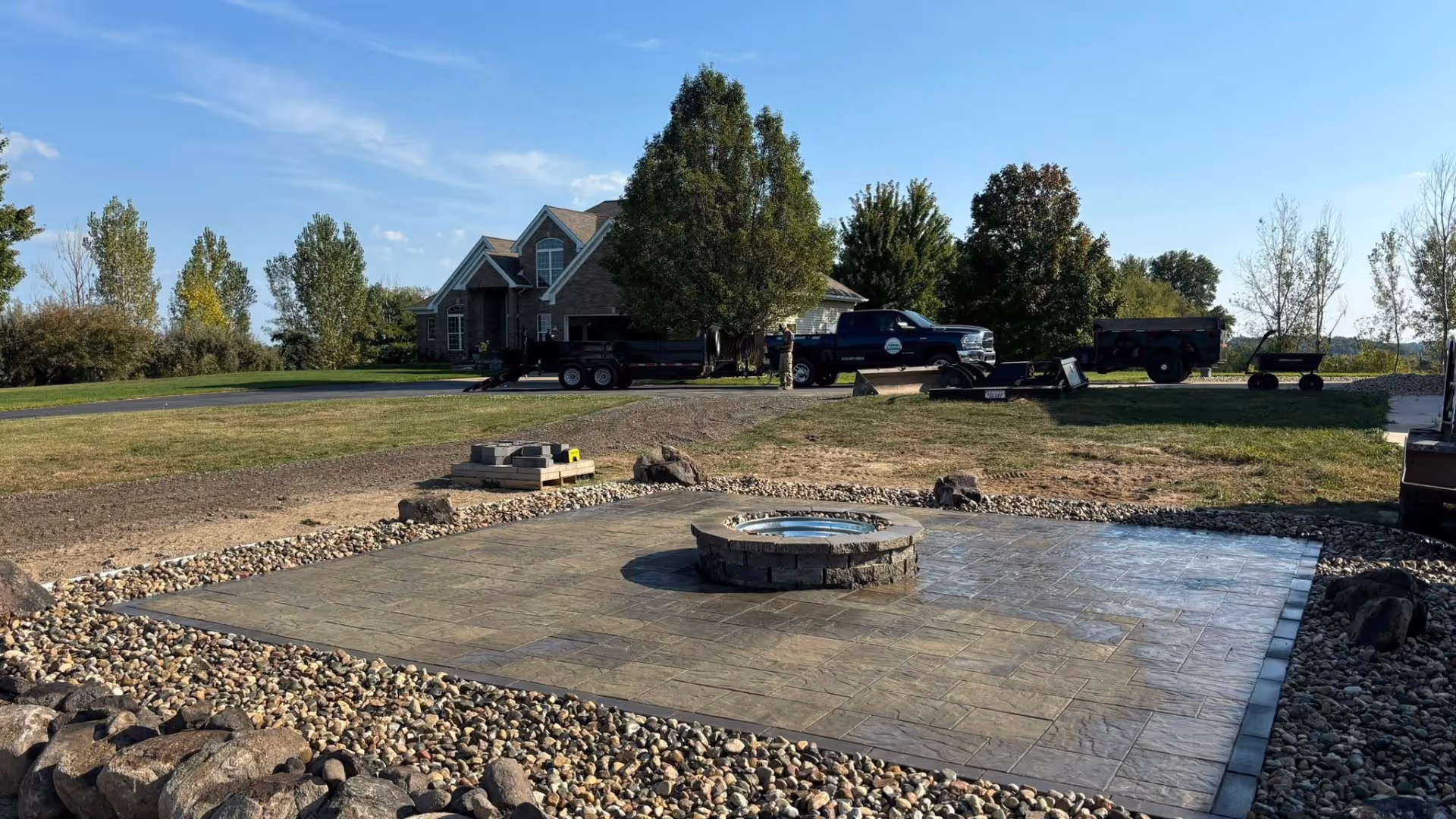 Stone-paved patio with a circular fire pit surrounded by pebbles in a backyard with trees and a house in the background.
