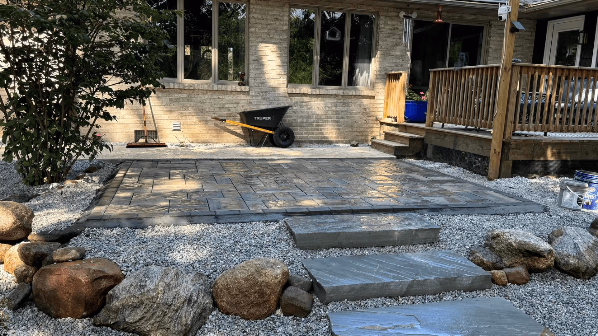 Patio area with rectangular stone tiles surrounded by gravel and large rocks, next to a wooden porch with steps and a wheelbarrow against a brick house wall.