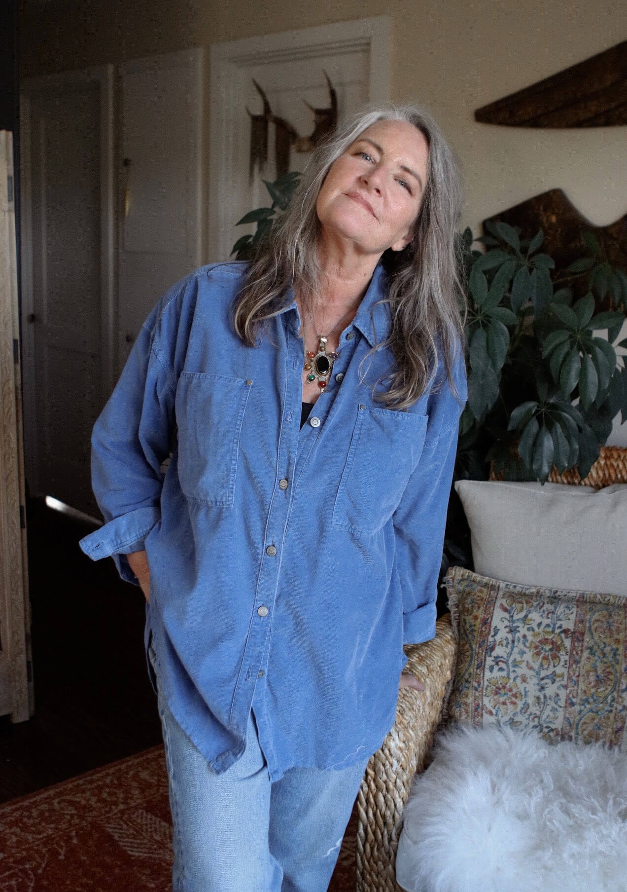 Smiling woman (Patty Davis) with long gray hair wearing a blue button-up shirt standing indoors near a wicker chair with cushions and a plant behind her.