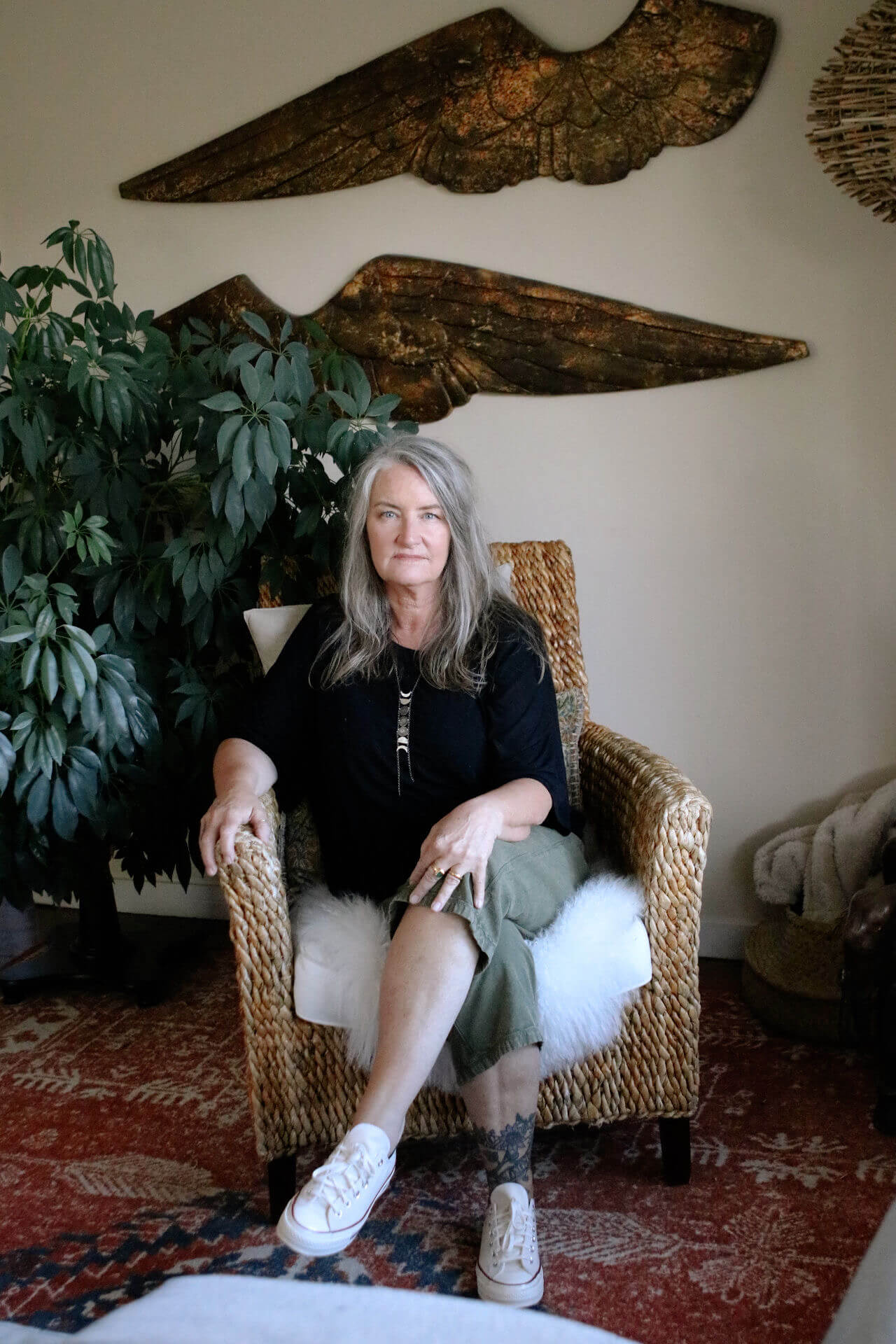Woman (Patty Davis) with long gray hair in black shirt and green pants sitting on a woven armchair with a plant and wooden wall art behind her.