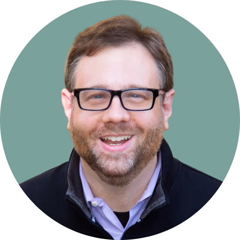 Smiling man with glasses, brown hair, and a beard wearing a purple shirt and black jacket.