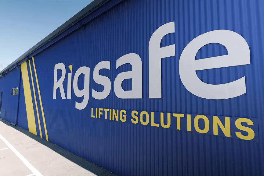 Blue corrugated metal building wall with the white and yellow logo text 'Rigsafe Lifting Solutions'.