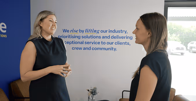 Two women in black dresses smiling and talking in an office with a motivational wall text about lifting the industry and delivering exceptional service.