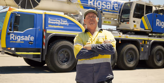 Man in yellow and navy high-visibility workwear standing confidently with crossed arms in front of a Rigsafe branded heavy lifting truck.