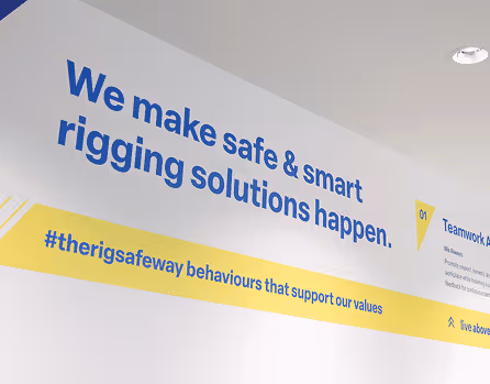 Wall text stating 'We make safe & smart rigging solutions happen.' with hashtag '#therigsafeway behaviours that support our values' on a yellow strip.