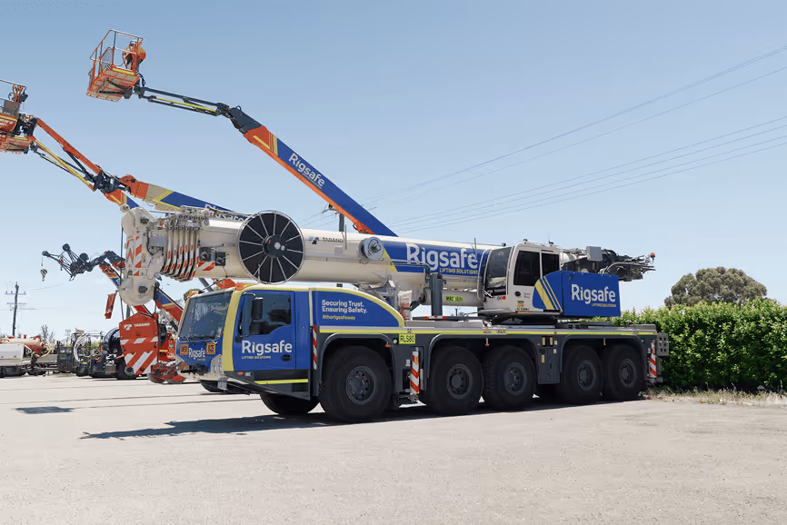 Large blue and white Rigsafe mobile crane with extended boom arm parked on a paved surface under a clear sky.