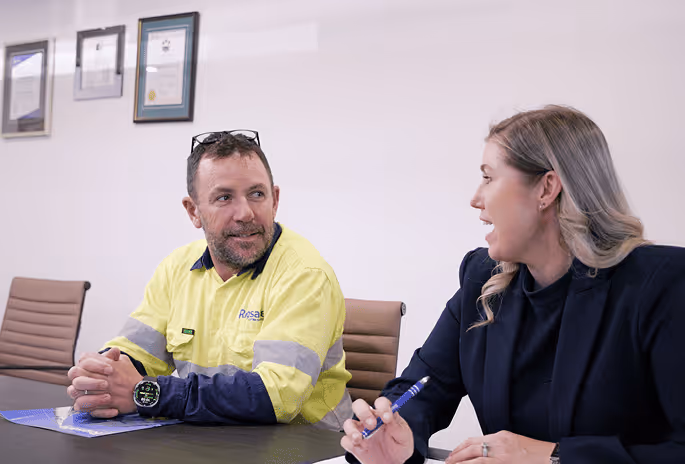 Man in a high-visibility work shirt and woman in business attire seated at a conference table engaged in conversation.