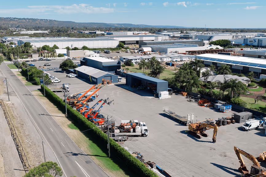Aerial view of an industrial yard with construction equipment and vehicles parked next to a long blue building under a partly cloudy sky.
