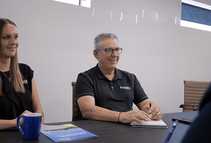 Two people sitting at a conference table, one man smiling and holding a pen with a notebook and a brochure on the table.