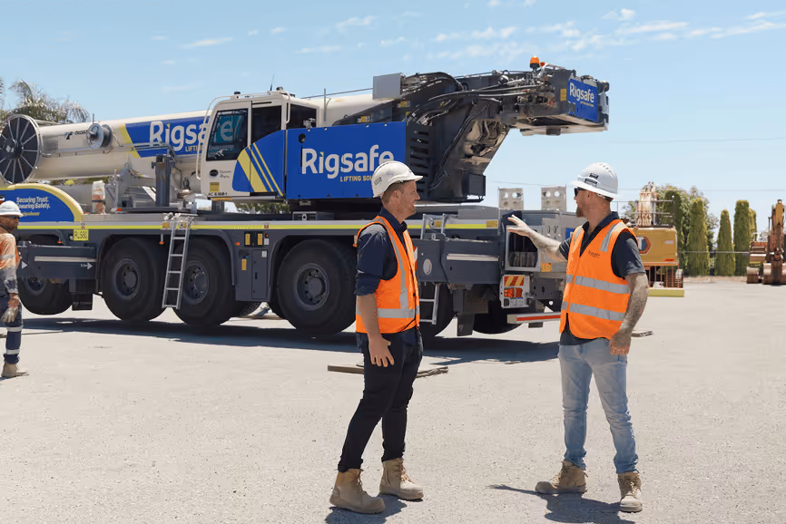 Two construction workers wearing orange safety vests and white helmets talking in front of a large Rigsafe crane truck in an outdoor work site.