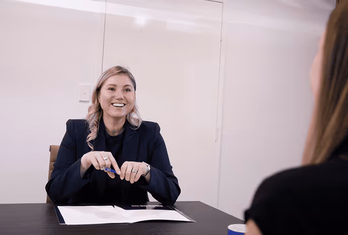A smiling woman in a black blazer holding a pen and sitting at a table with documents during a conversation with another person.