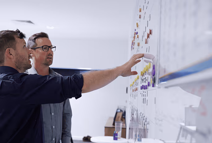 Two men discussing and pointing at notes and magnets on a whiteboard in an office.