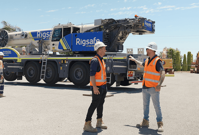 Two construction workers wearing white helmets and orange safety vests talking in front of a large Rigsafe crane truck outdoors.